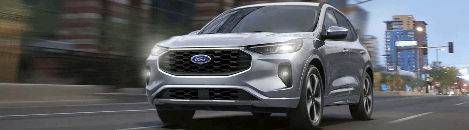 A silver SUV with a prominent grille and headlights parked on a city street, with blurred buildings and structures visible in the background.