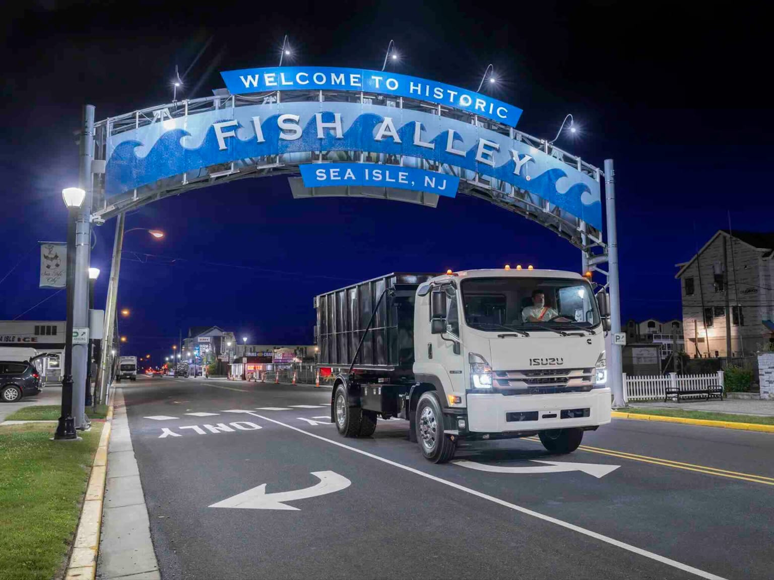 A white truck drives under a brightly lit archway at night, with a street and buildings visible in the background.