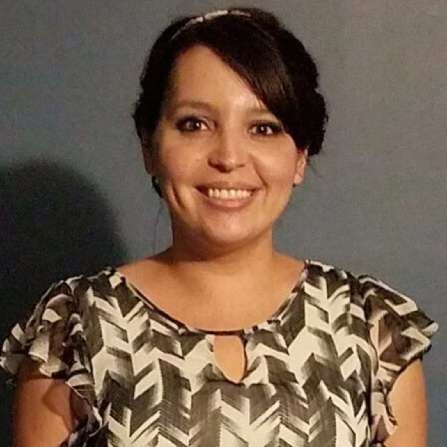 A smiling woman with dark hair wearing a patterned blouse stands in front of a plain background.