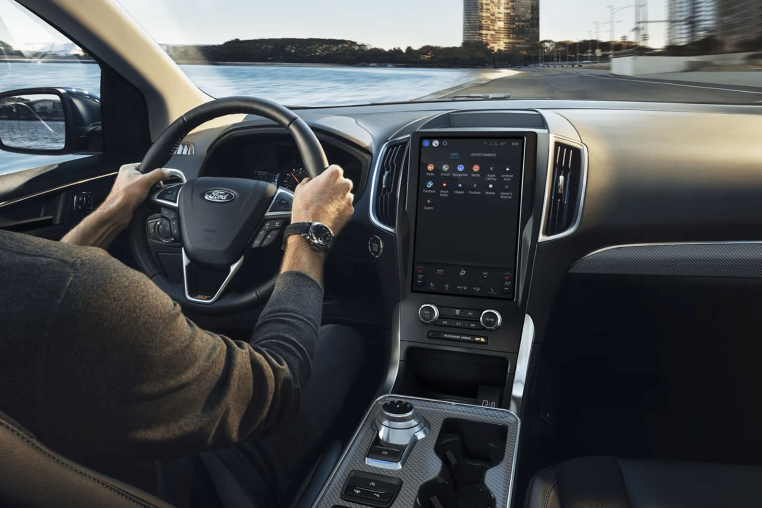 The image shows the interior of a car, with the driver's hands on the steering wheel and the dashboard displaying various controls and a large touchscreen display. In the background, a cityscape with tall buildings can be seen through the windshield.