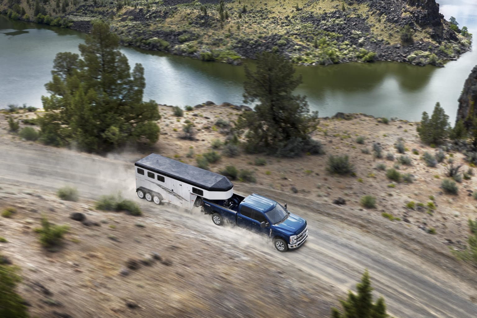 a blue truck driving down a dirt road next to a lake and a mountain covered in trees and shrubs