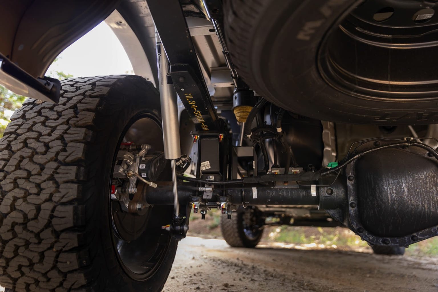 The image depicts the undercarriage of a vehicle with a large off-road tire in the foreground and various mechanical components and suspension parts visible in the background.