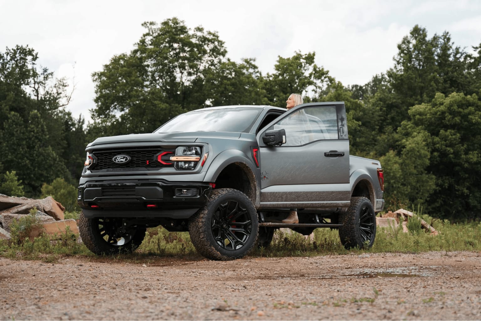 A rugged, lifted truck stands on a dirt road surrounded by lush, green trees in the background.