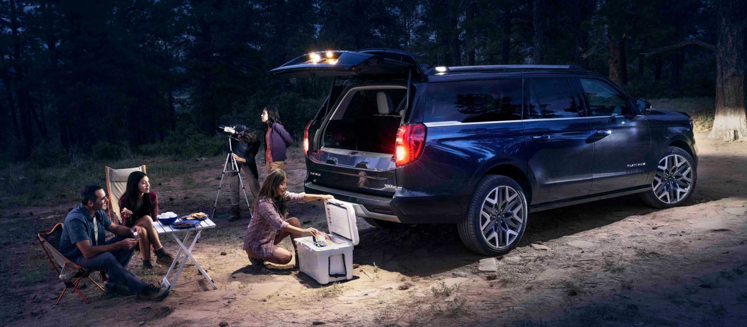Dark blue SUV in a forest with people sitting around a campfire
