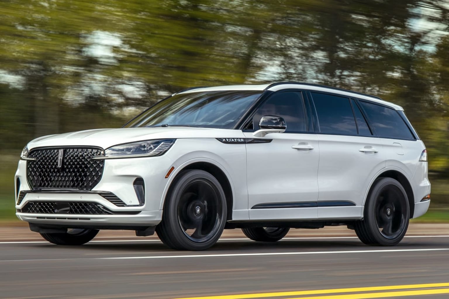 A white SUV with a sleek, modern design is speeding down a road surrounded by a blurred, forested landscape.