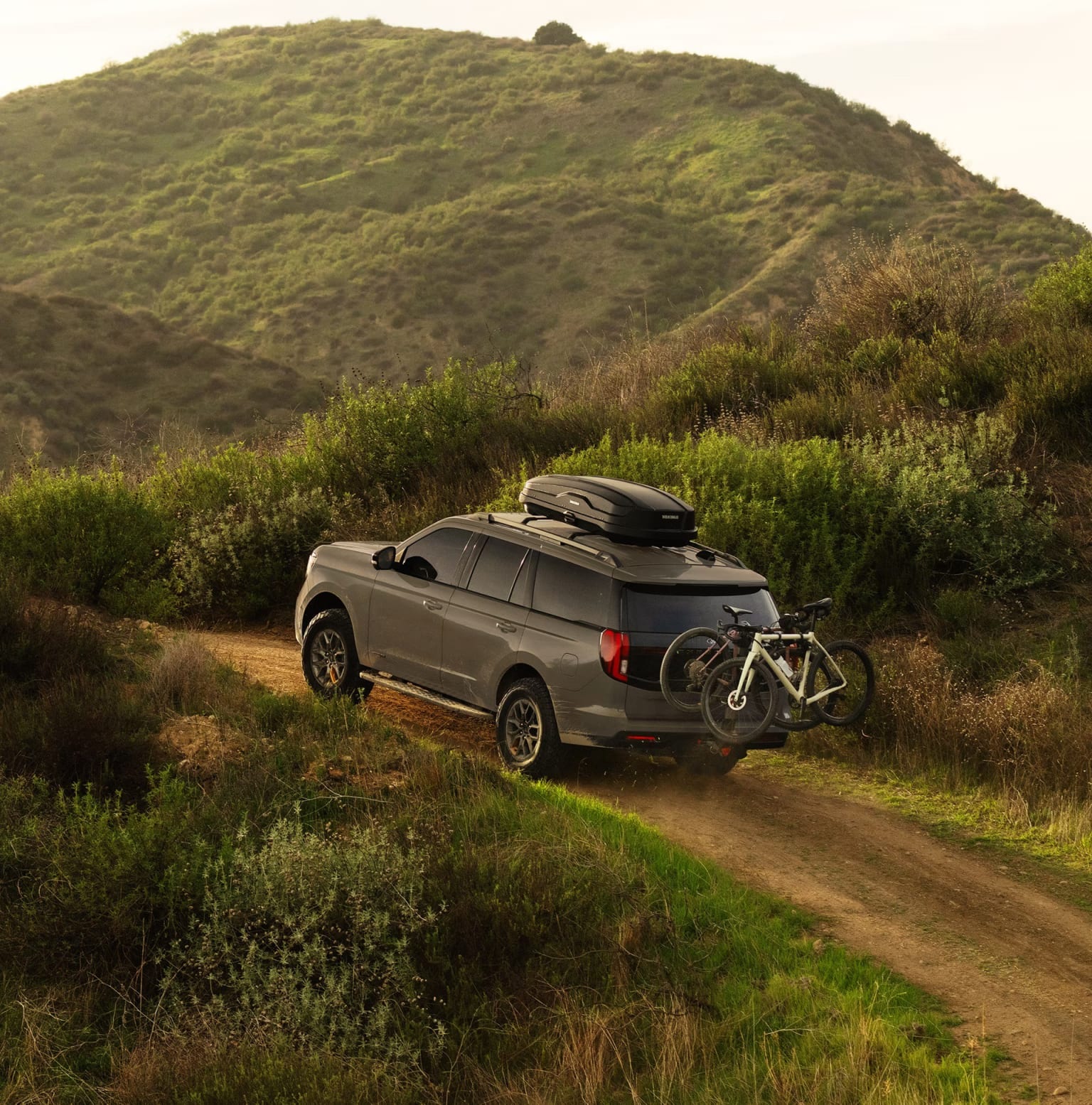 A gray SUV with a bike rack and a roof box drives on a dirt road through a green, hilly landscape.