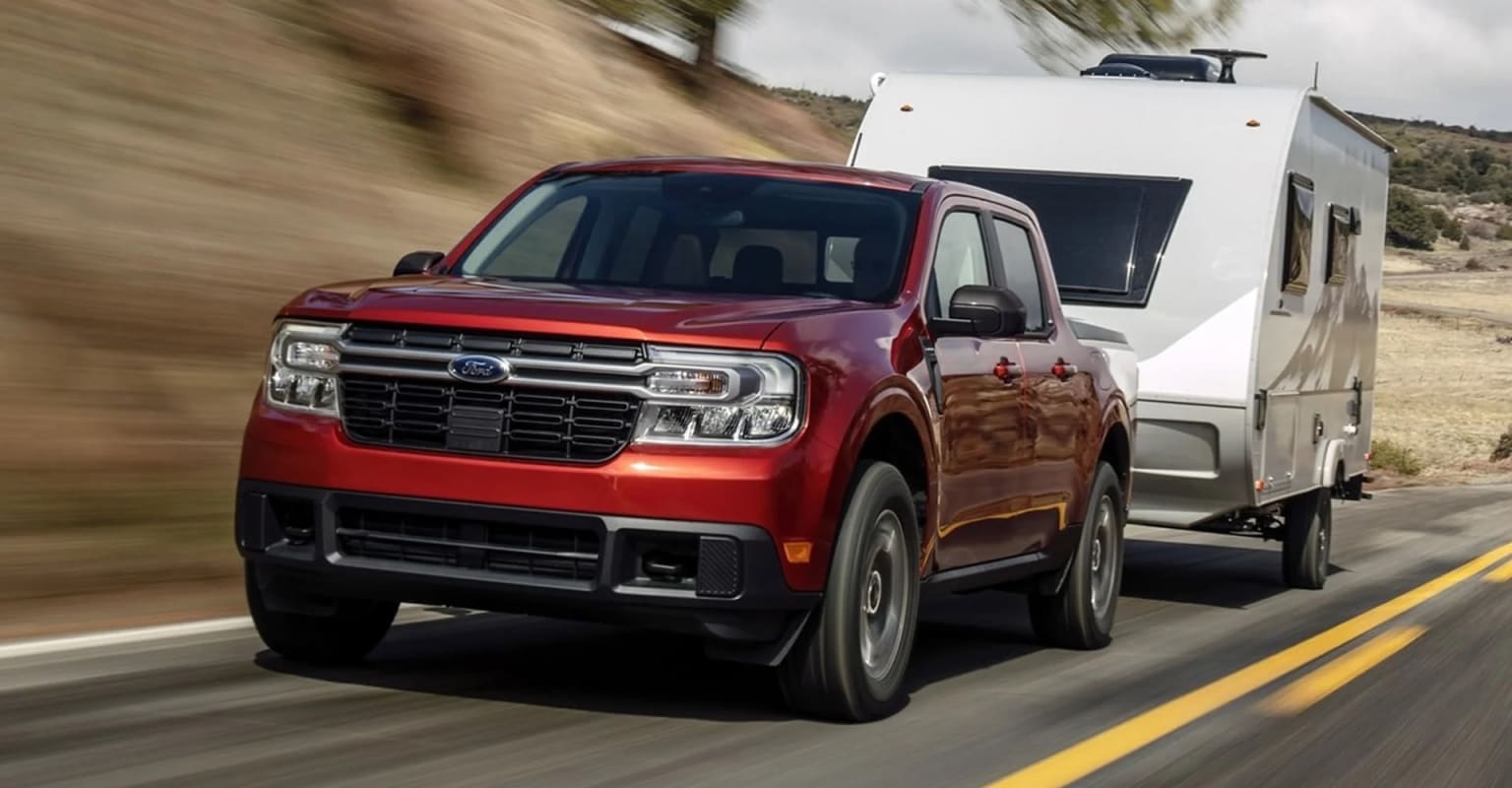 A red pickup truck towing a white camper trailer is speeding down a winding road surrounded by a mountainous, forested landscape.