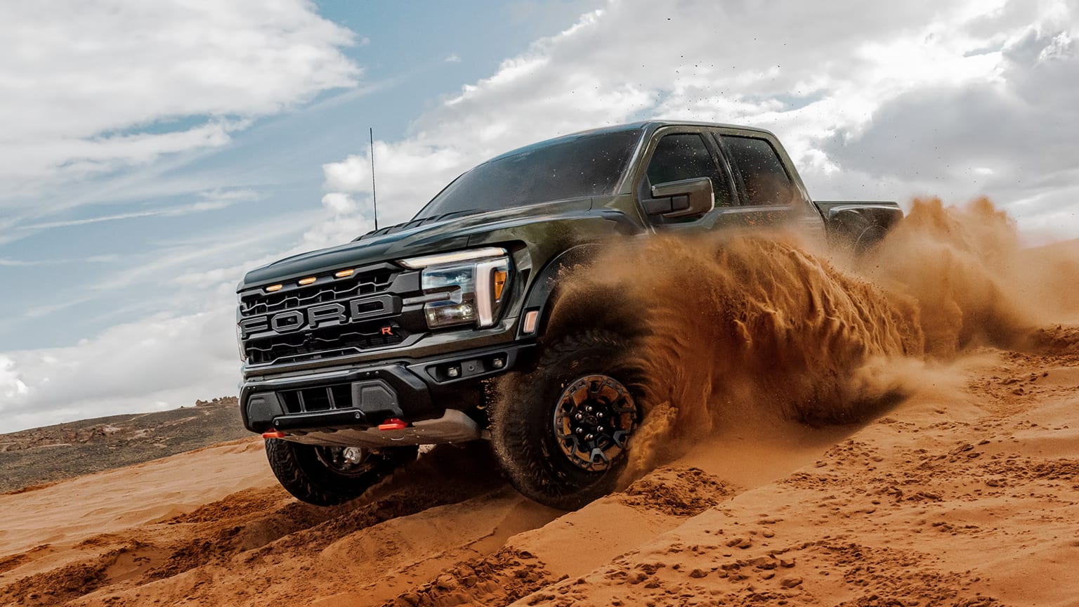 A dark gray pickup truck kicks up clouds of sand while driving across a desert dune under a cloudy sky.