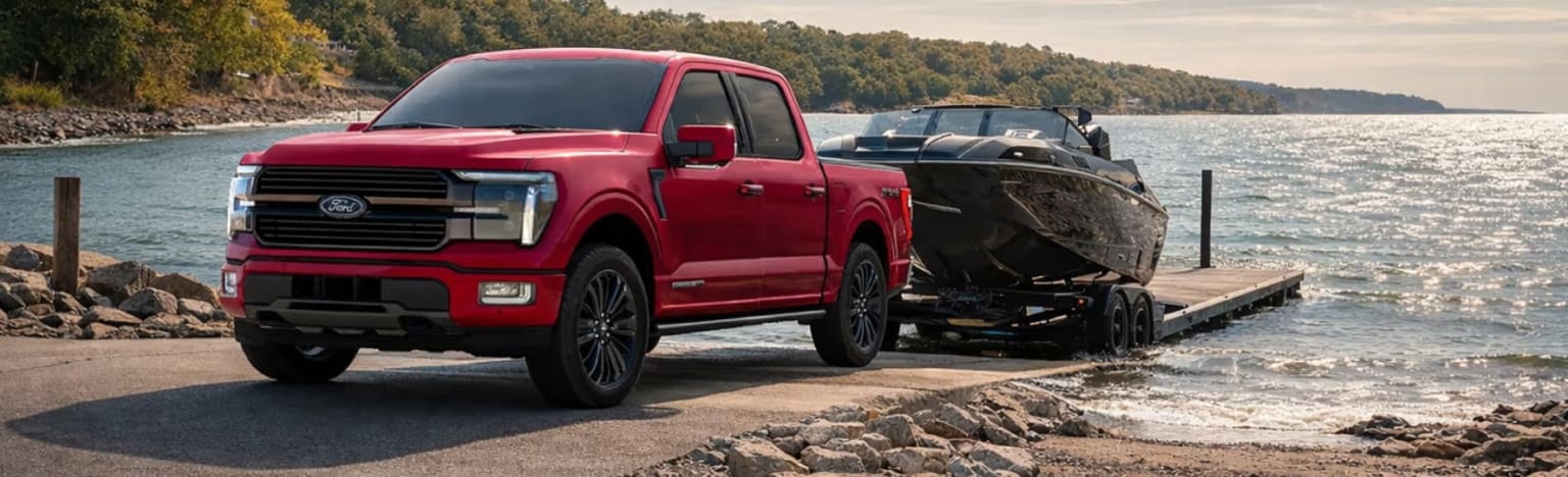 A red pickup truck towing a boat on a trailer down a ramp towards a sparkling lake, with a tree-lined shore in the background.