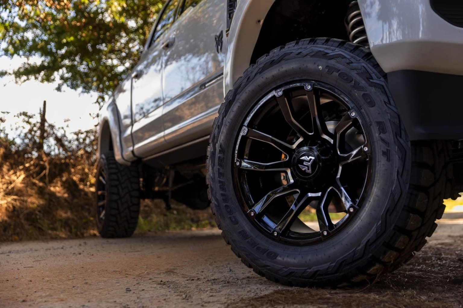 The image shows a close-up view of a large off-road vehicle tire with a rugged tread pattern, set against a backdrop of a dirt road and lush foliage in the background.