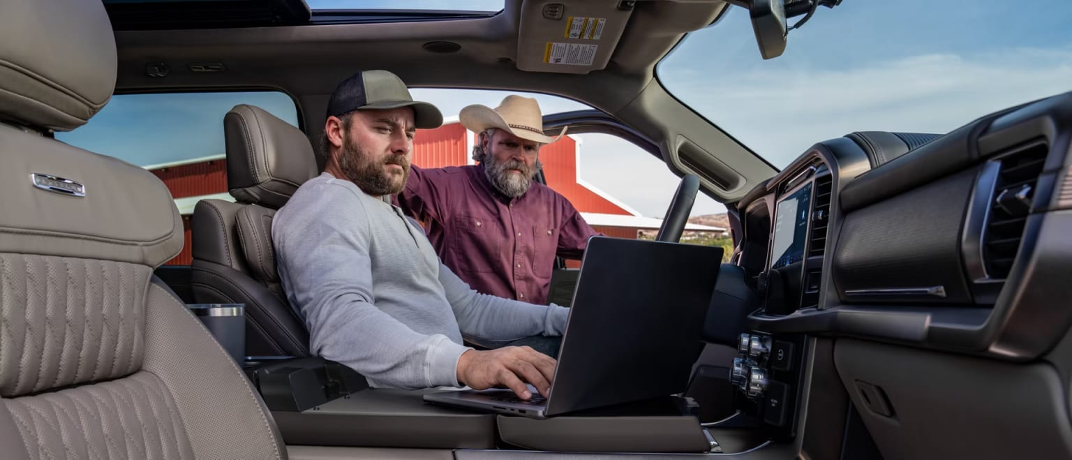Two men in a truck cab, one using a laptop, with a red barn and blue sky in the background.