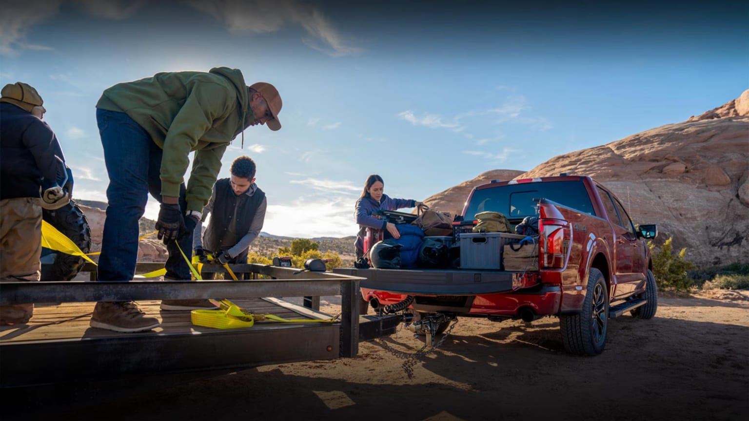 A group of people gathered around an off-road vehicle in a desert landscape, with rocky formations and a blue sky in the background.