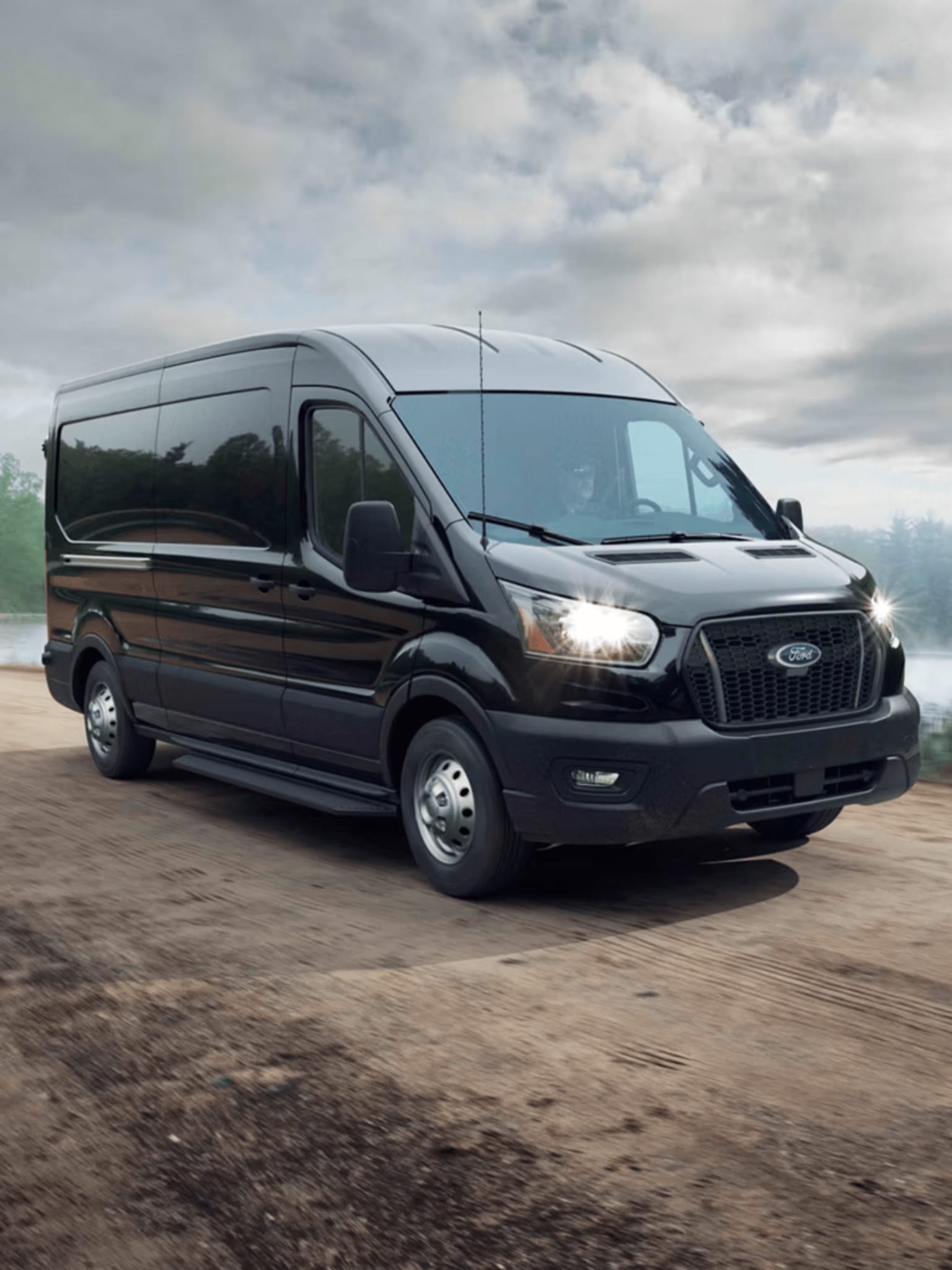 A black van is parked on a dirt road with a cloudy sky and trees in the background.