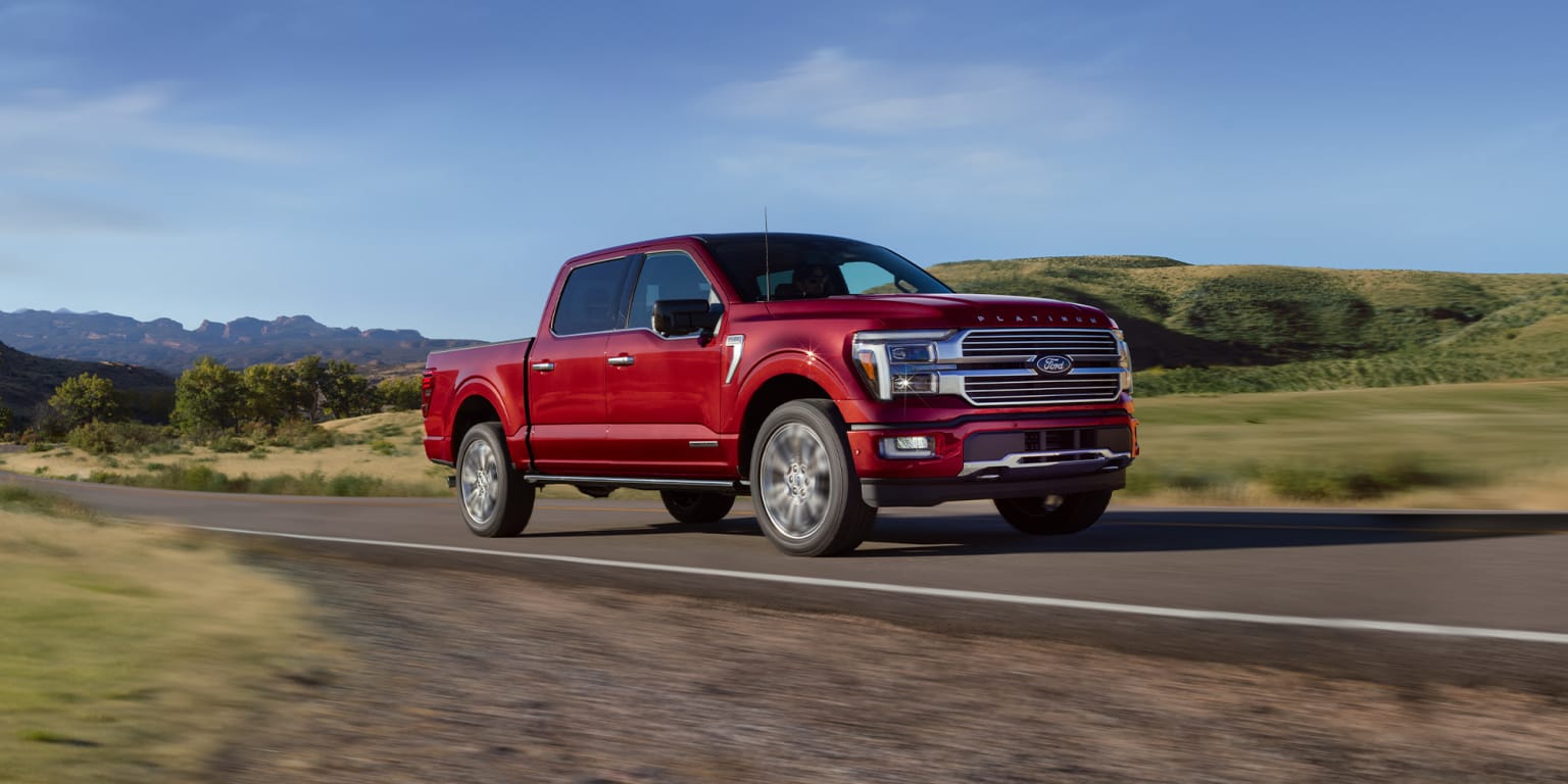 A red pickup truck driving on a road with hills and vegetation in the background.