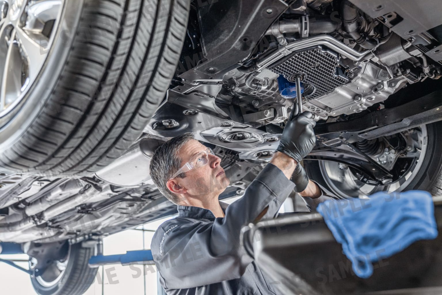 A mechanic in a blue uniform conducting maintenance on the underside of a car, inspecting the engine components.