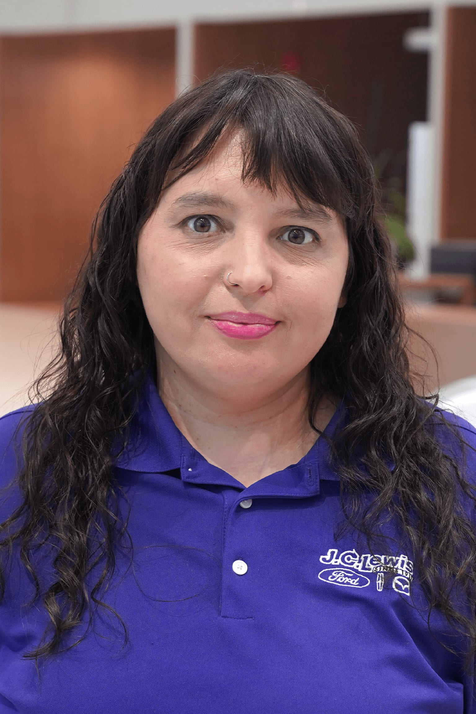 A woman with curly dark hair in a blue polo shirt standing in an indoor setting with a wooden background.