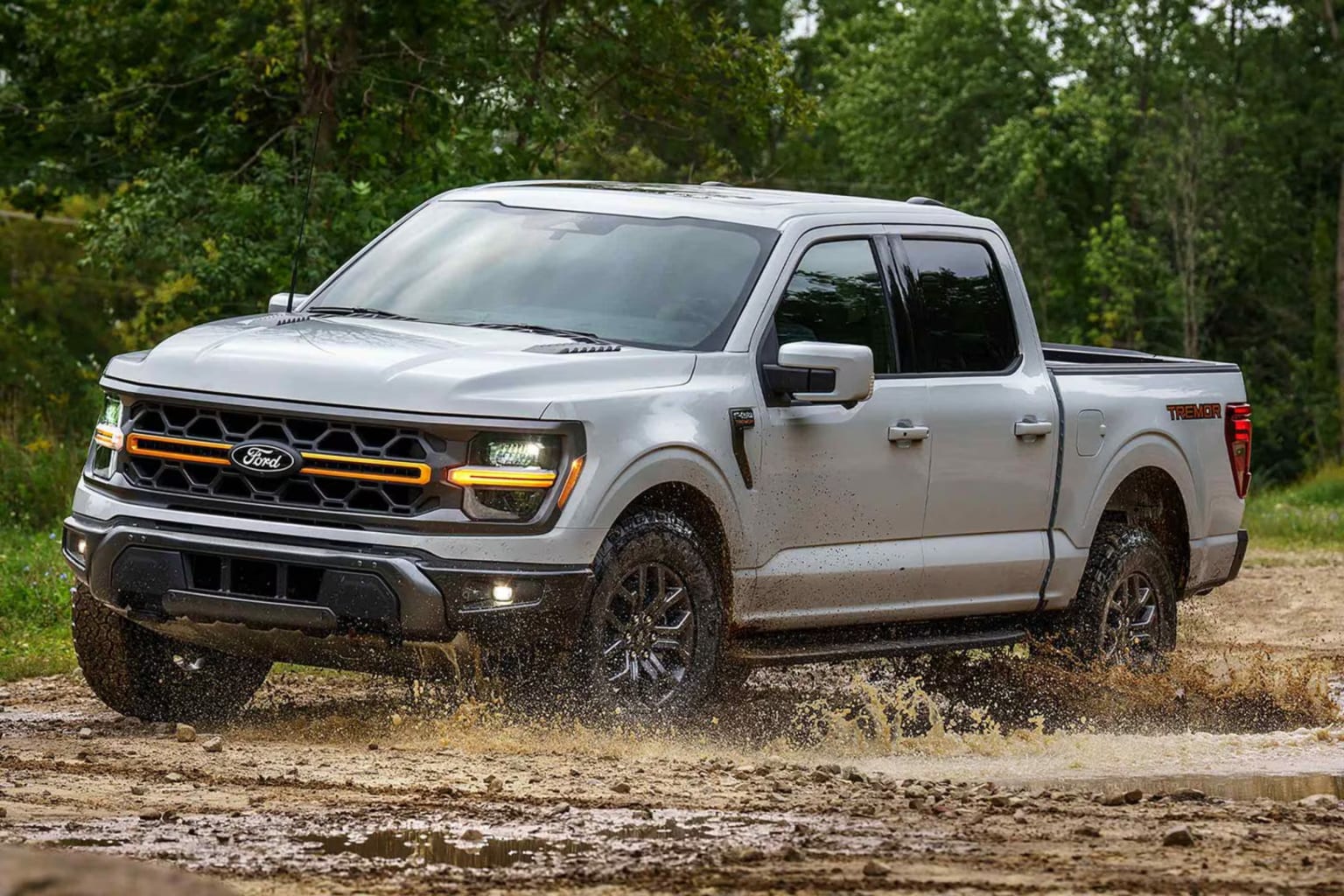 A silver pickup truck driving through a muddy puddle with a blurred green forest in the background.