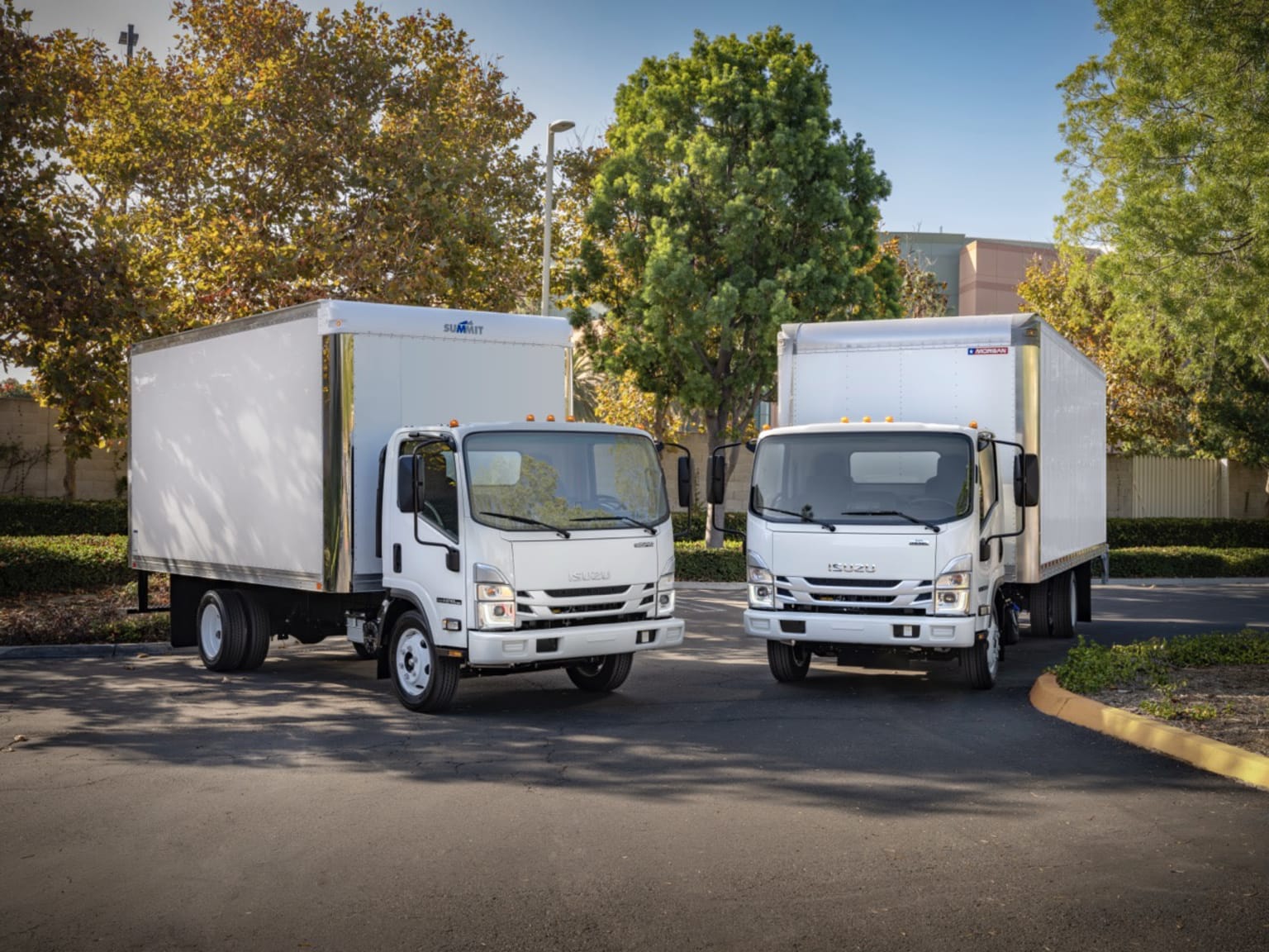 Two white box trucks on an asphalt lot with trees and a building in the background.