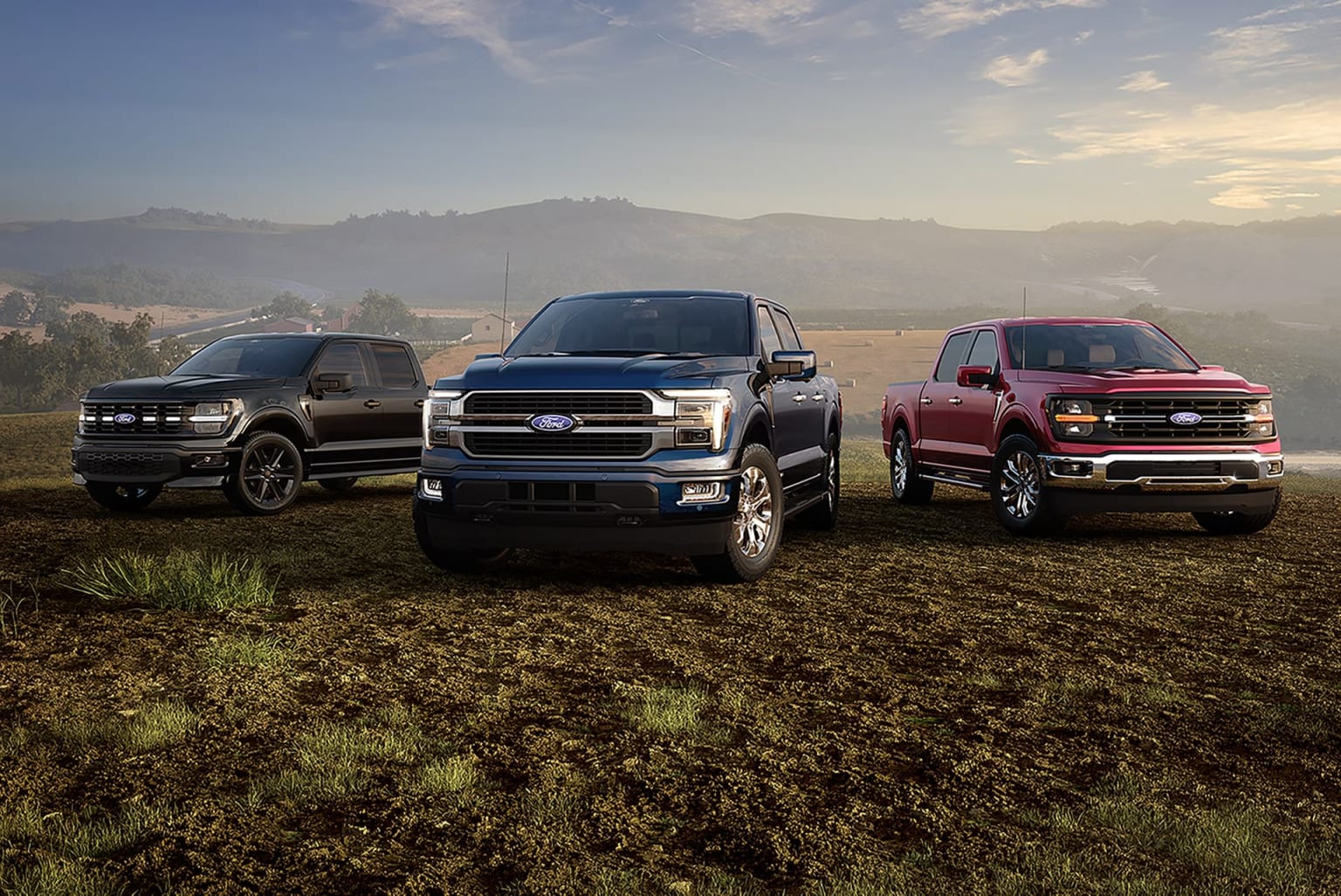 Three pickup trucks in various colors parked on a grassy field with mountains visible in the background under a cloudy sky.