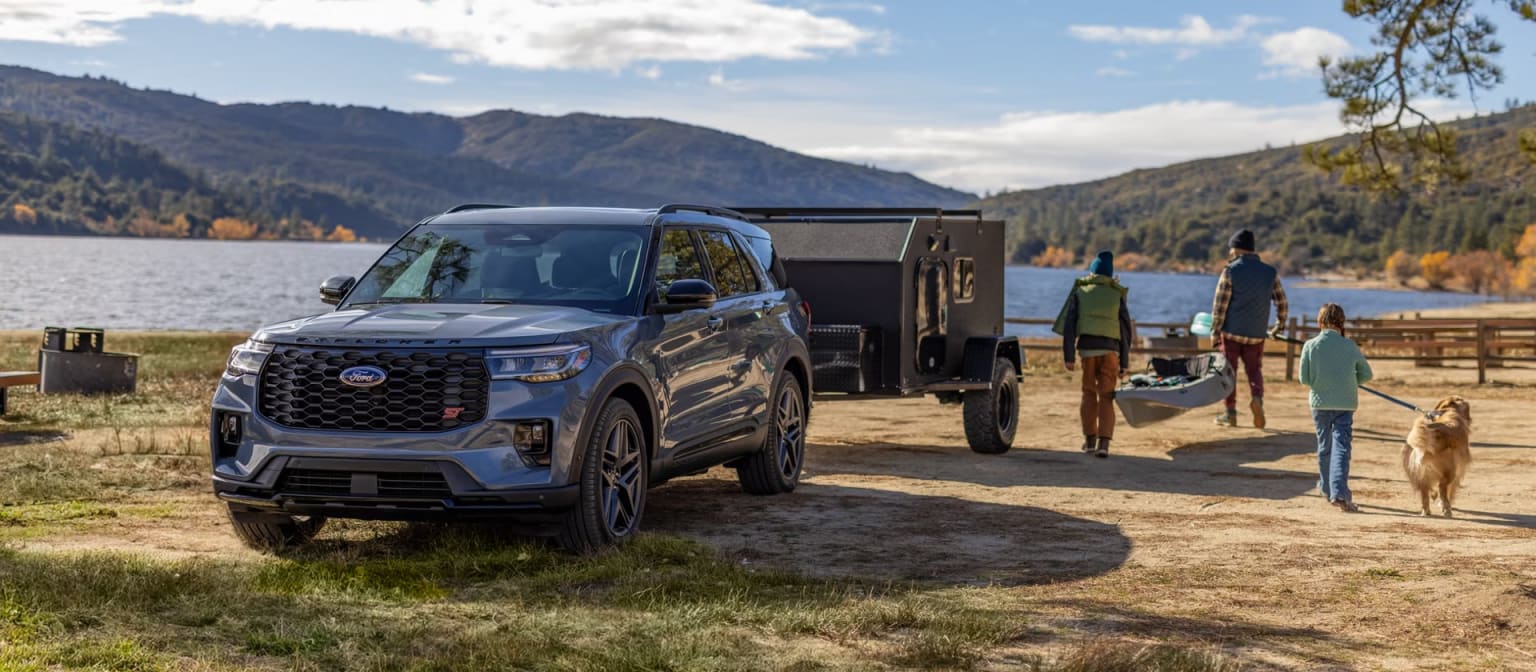 A large black SUV with a trailer is parked on a dirt road near a lake, surrounded by mountains and trees in the background.