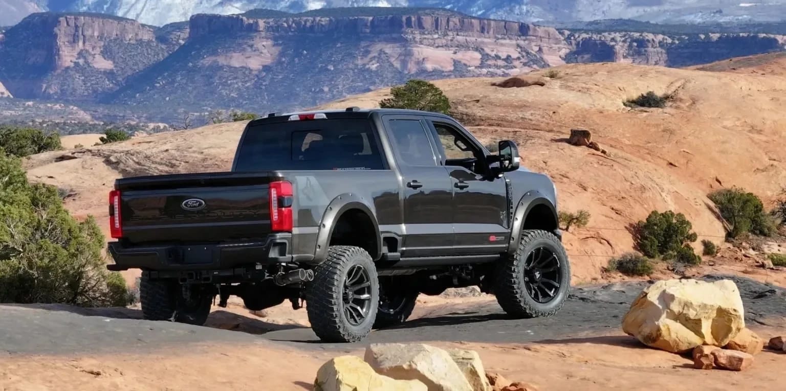 A dark gray truck with large tires is parked on a rocky, desert terrain with mesas and mountains in the background.