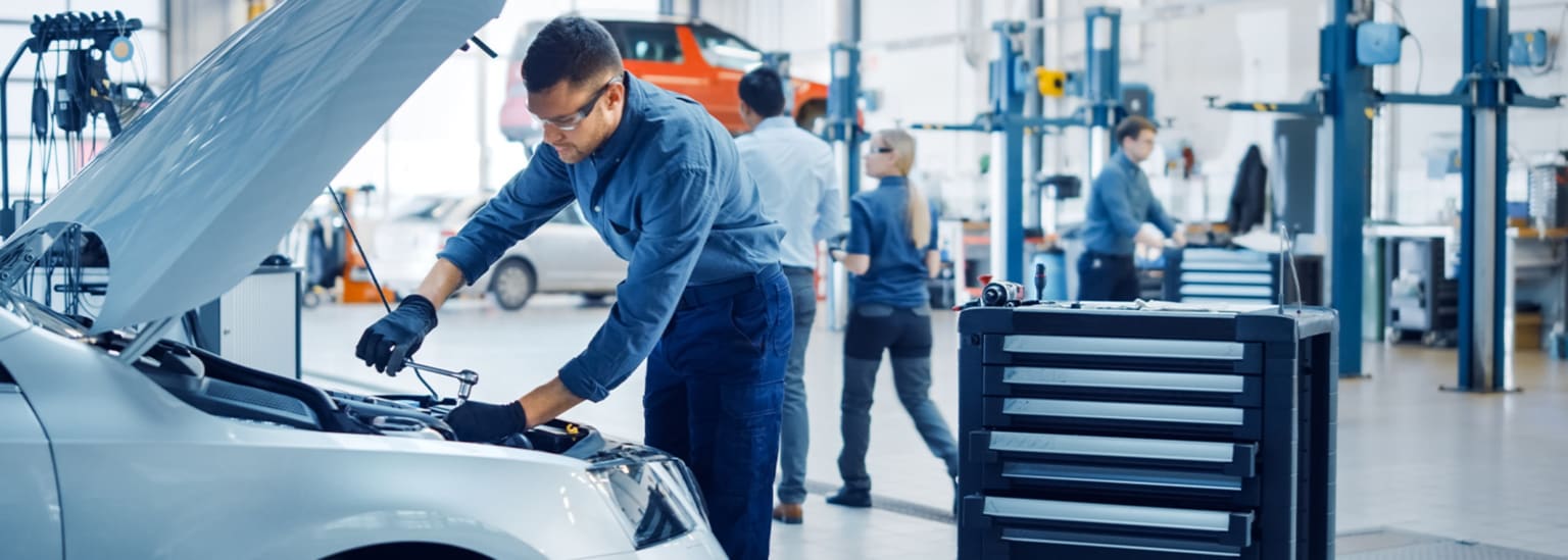An auto mechanic is working on a car's engine in a busy auto repair shop. Other people and car lifts are visible in the background.