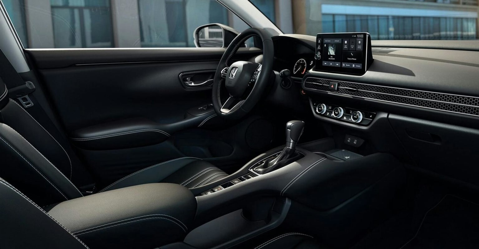 A modern car interior with a touchscreen display and dashboard in the foreground, and a blurred urban cityscape visible through the windows in the background.