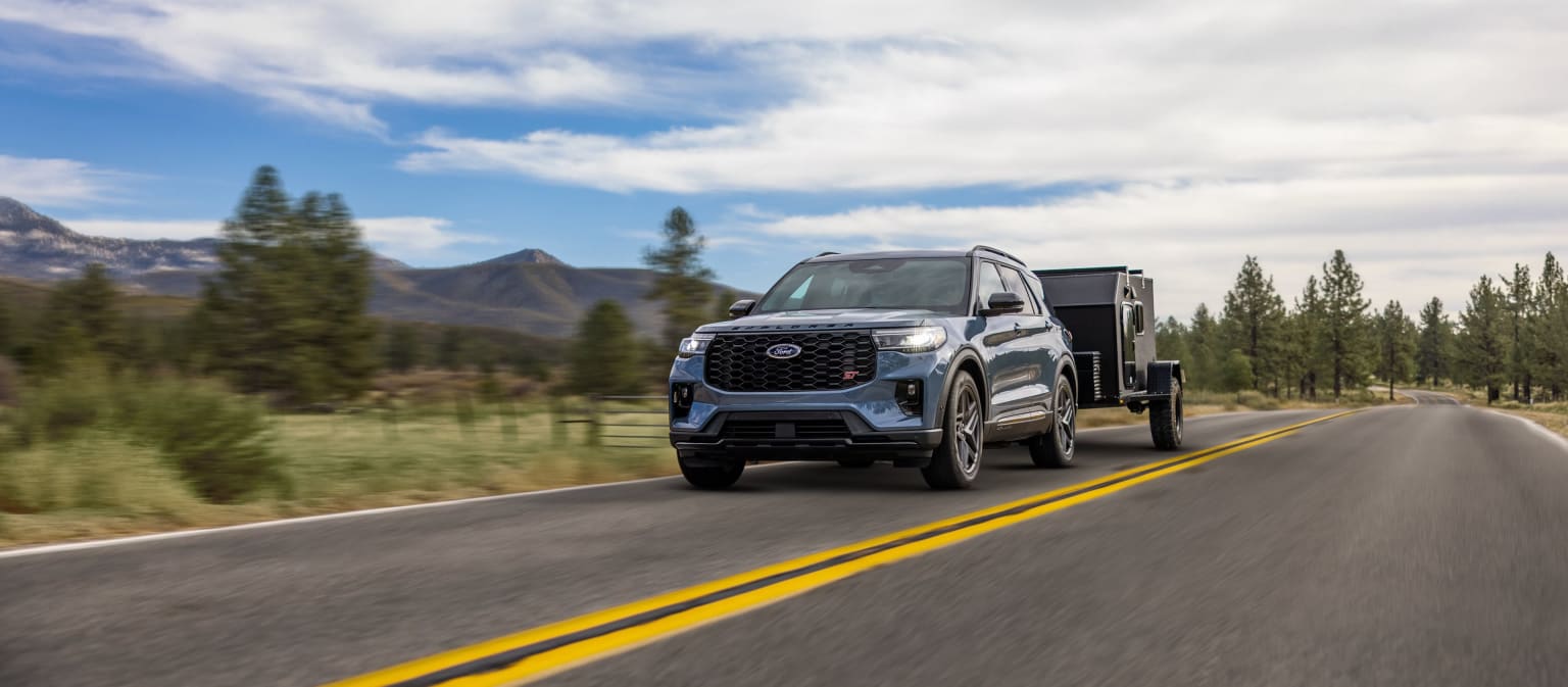 A large, rugged-looking pickup truck with a dump bed is driving down a winding mountain road, surrounded by pine trees and snow-capped peaks in the background.