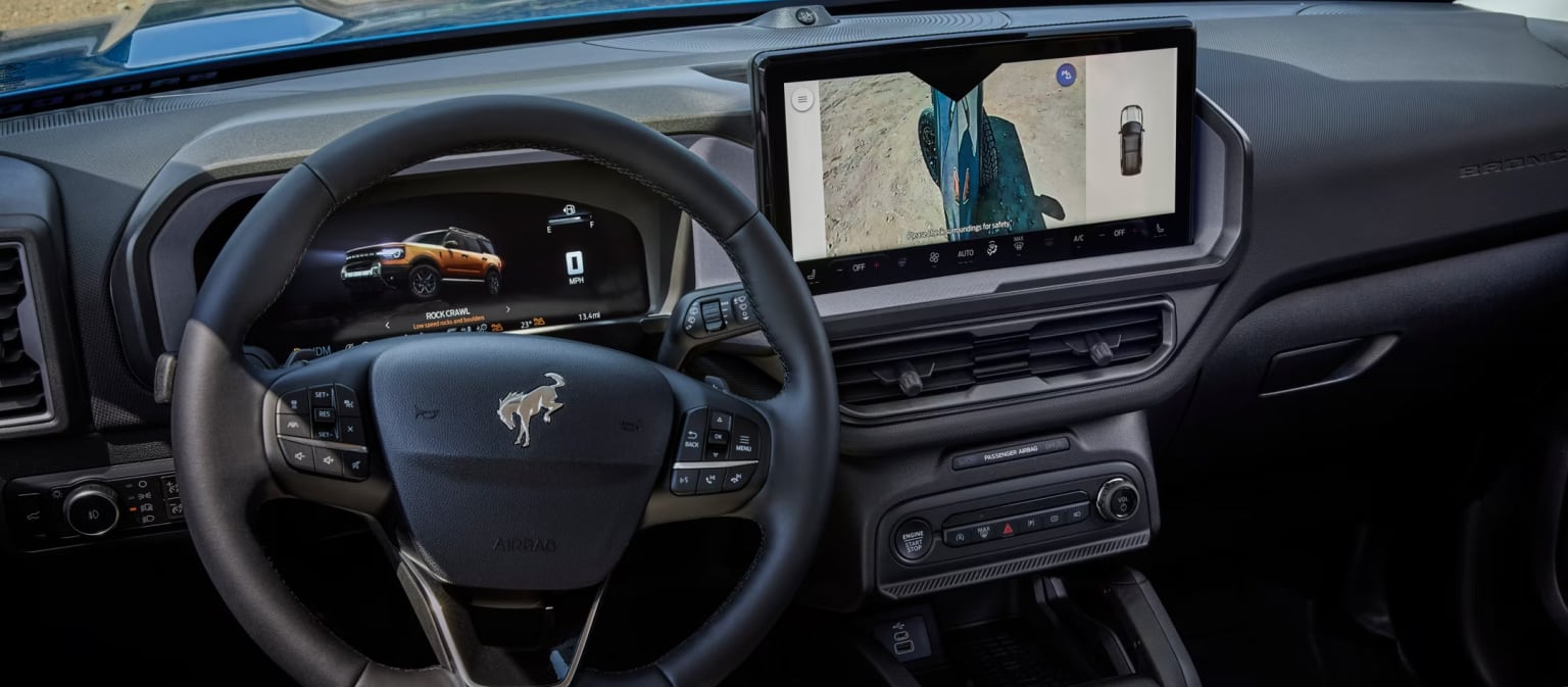 Interior of a vehicle with a large touchscreen display in the center console showing a map, along with various controls and buttons on the steering wheel and dashboard.