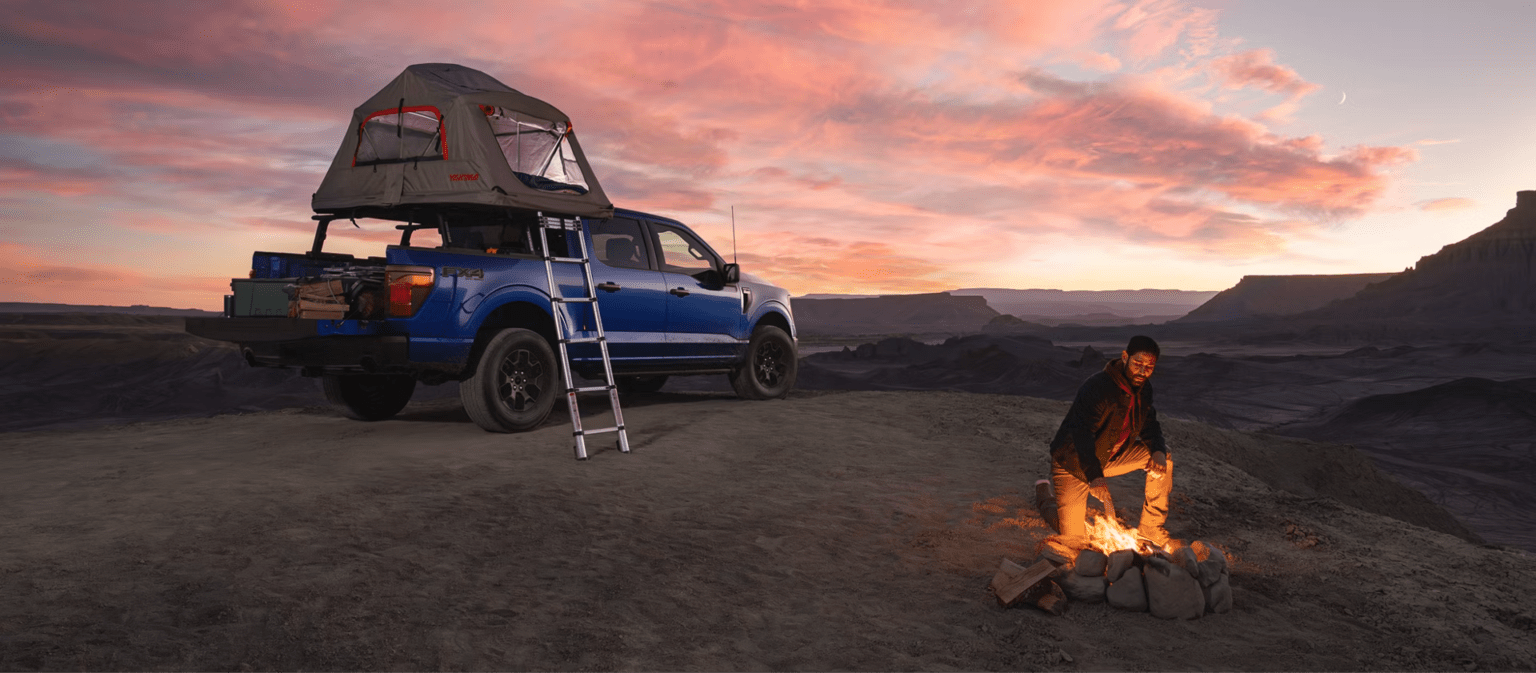 A man by a campfire with a blue pickup truck and rooftop tent parked in a desert landscape at sunset.