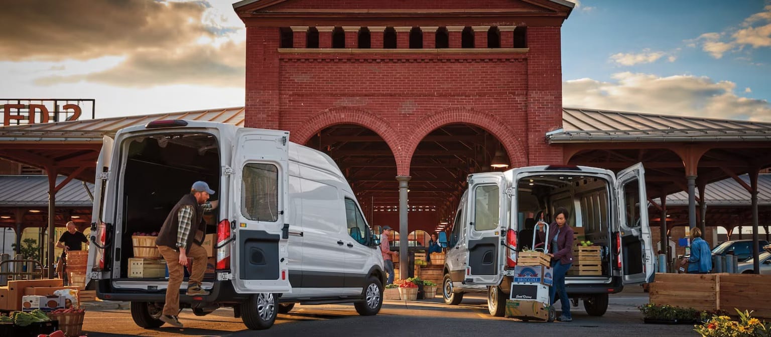 Two white delivery vans being unloaded by workers in front of a large red brick market building under a cloudy sky.