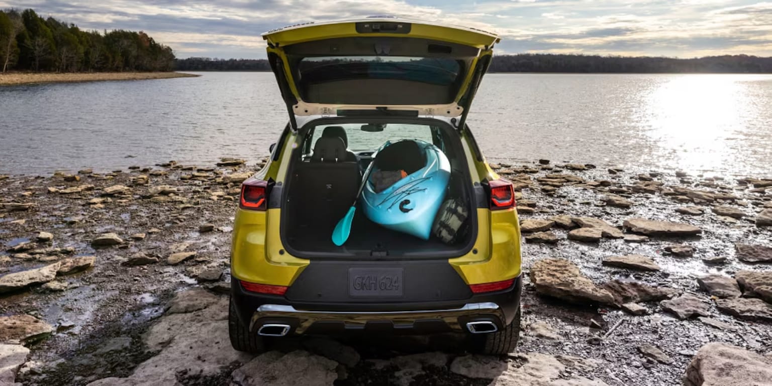 A yellow car parked on a rocky beach near a body of water, with its trunk open and a surfboard inside.