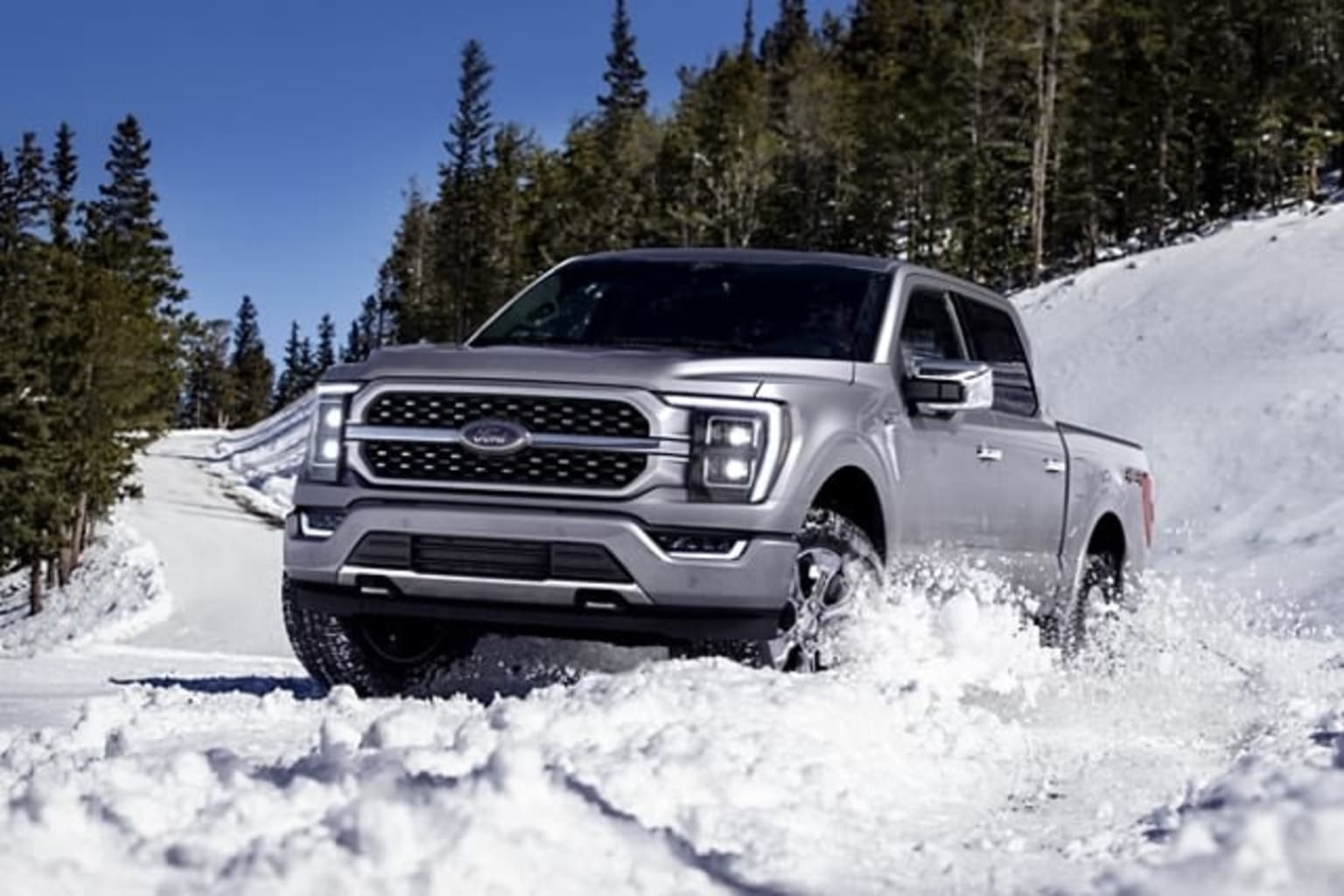 a silver truck driving down a snow covered road next to a pine tree covered hill in the background is a blue sky and white clouds