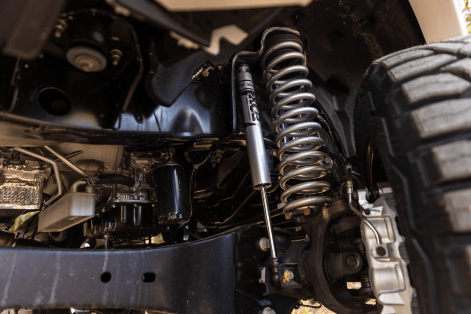 Coilover shock absorber and suspension components displayed in silver color in the foreground, with the underside of a vehicle and a tire in the background.