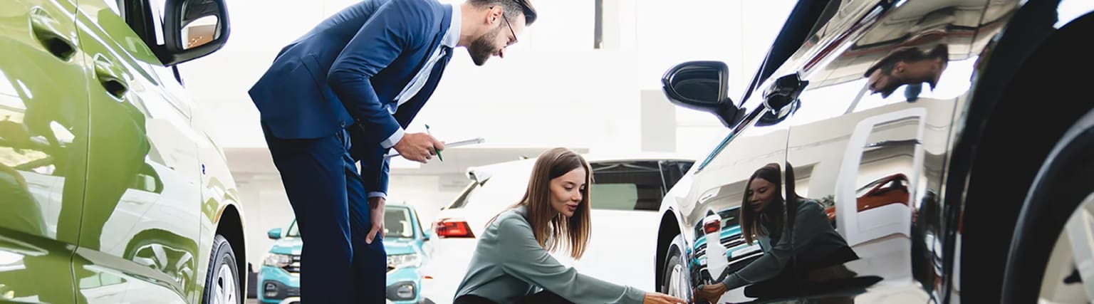 A man in a suit is examining a car while a woman in casual clothing appears to be observing him, with another person visible in the background.