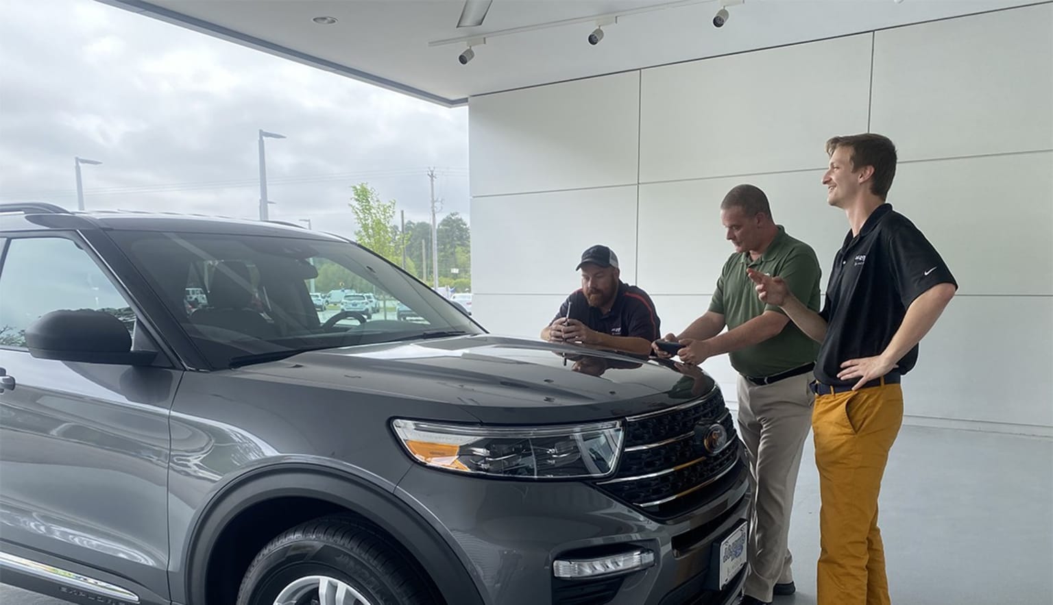 a couple of men standing next to a gray car in a showroom next to another man pointing at the hood of a car