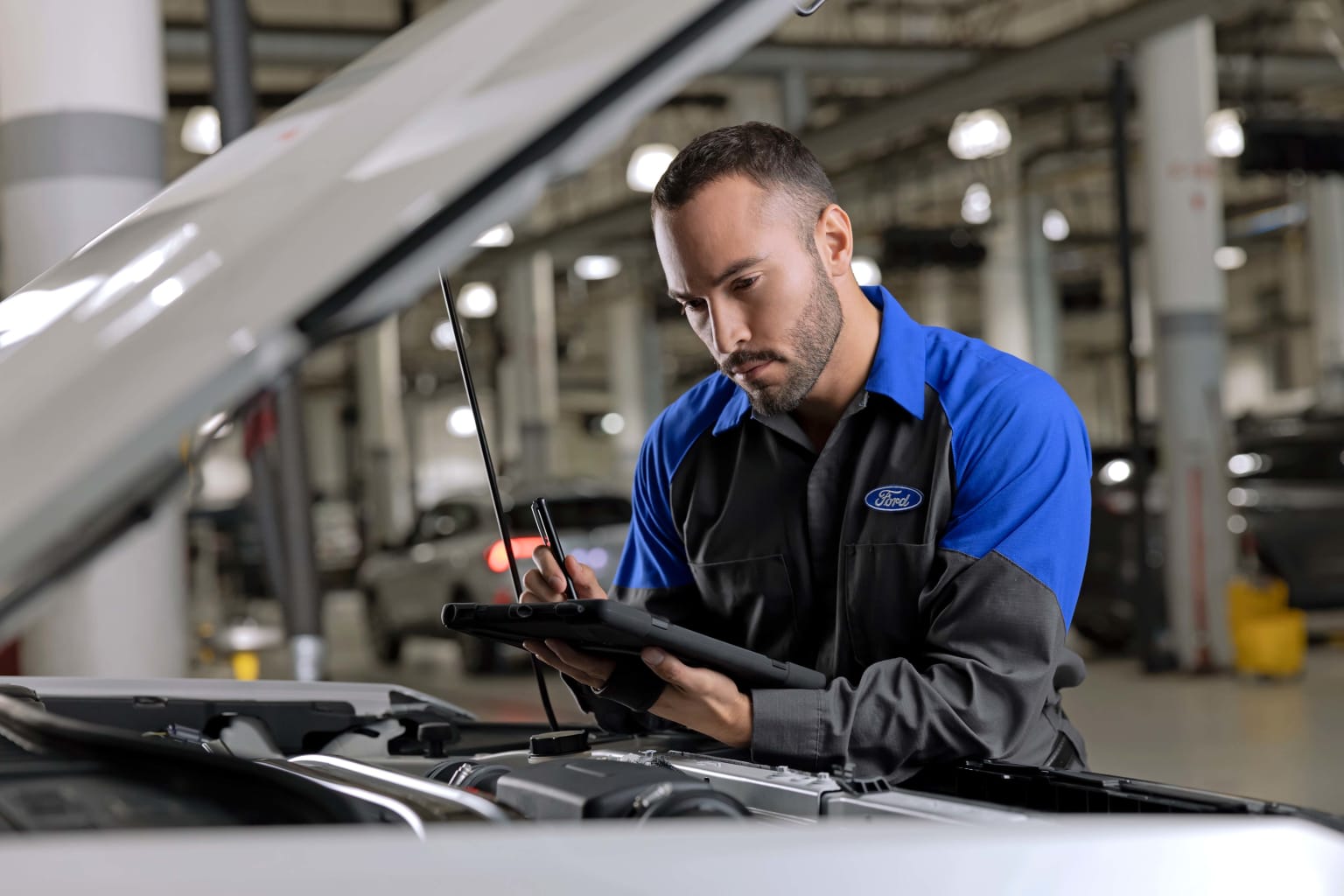 a man in a blue shirt working on a car's hood in a garage with a wrench in his hand