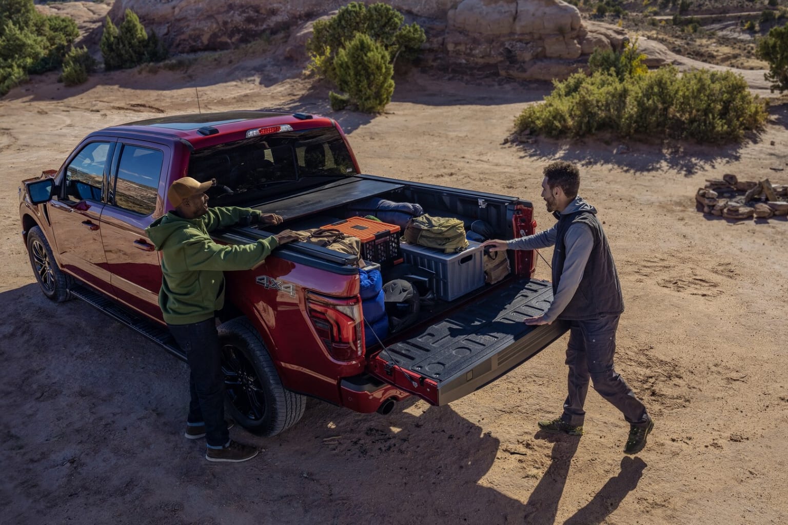 Image of a red pickup truck parked in a desert-like landscape with a person next to the open truck bed filled with camping and outdoor gear.