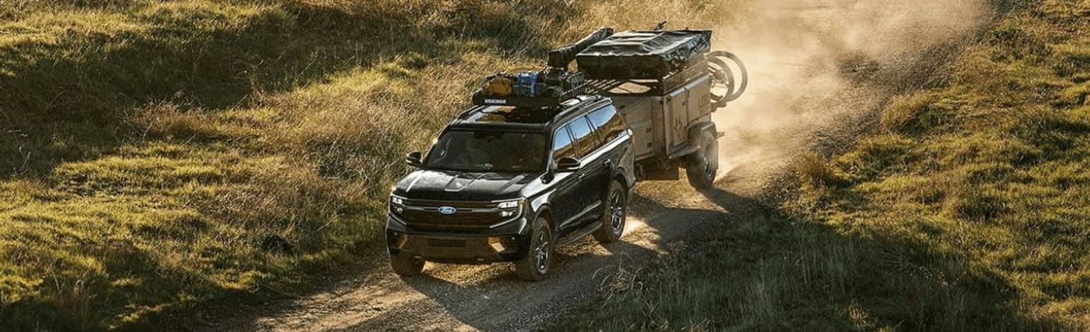 Black SUV towing a trailer on a dusty dirt road through a grassy, hilly landscape.