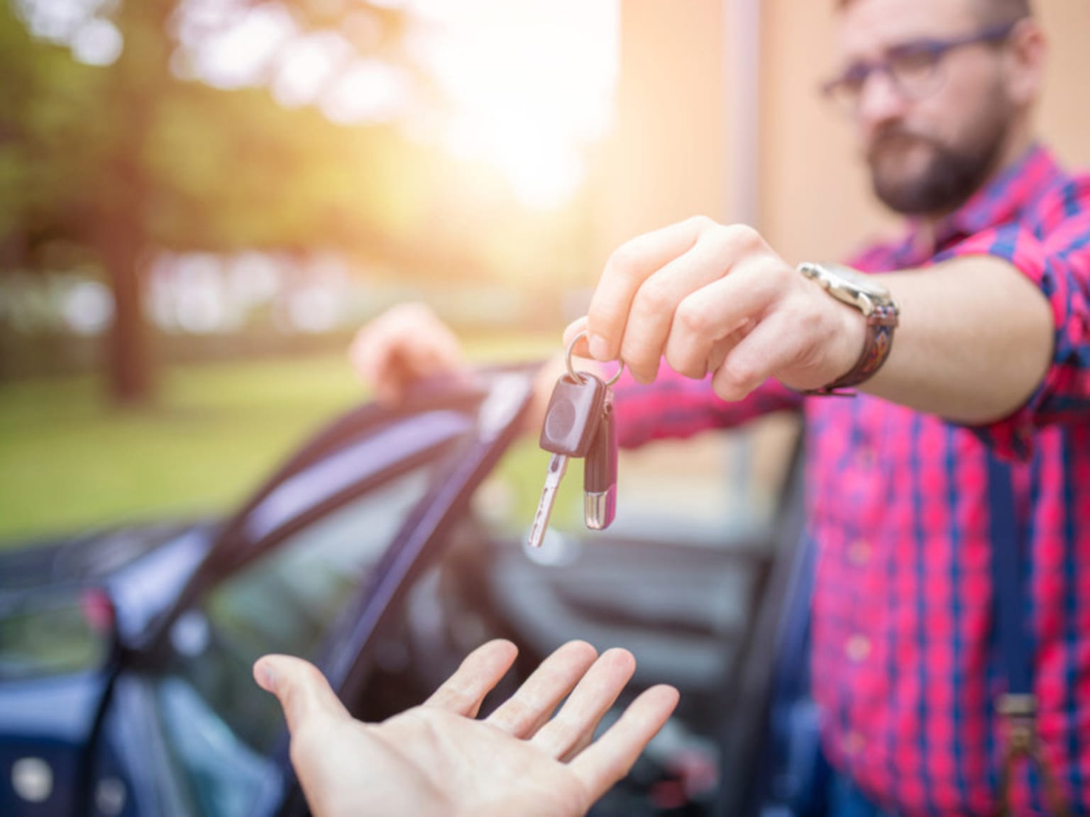 A person's hand holding car keys in the foreground, with a blurred background of a vehicle and a sunny outdoor setting.