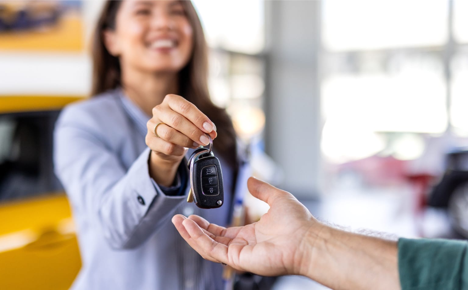A smiling woman handing car keys to a man, with a blurred yellow car and dealership interior in the background.