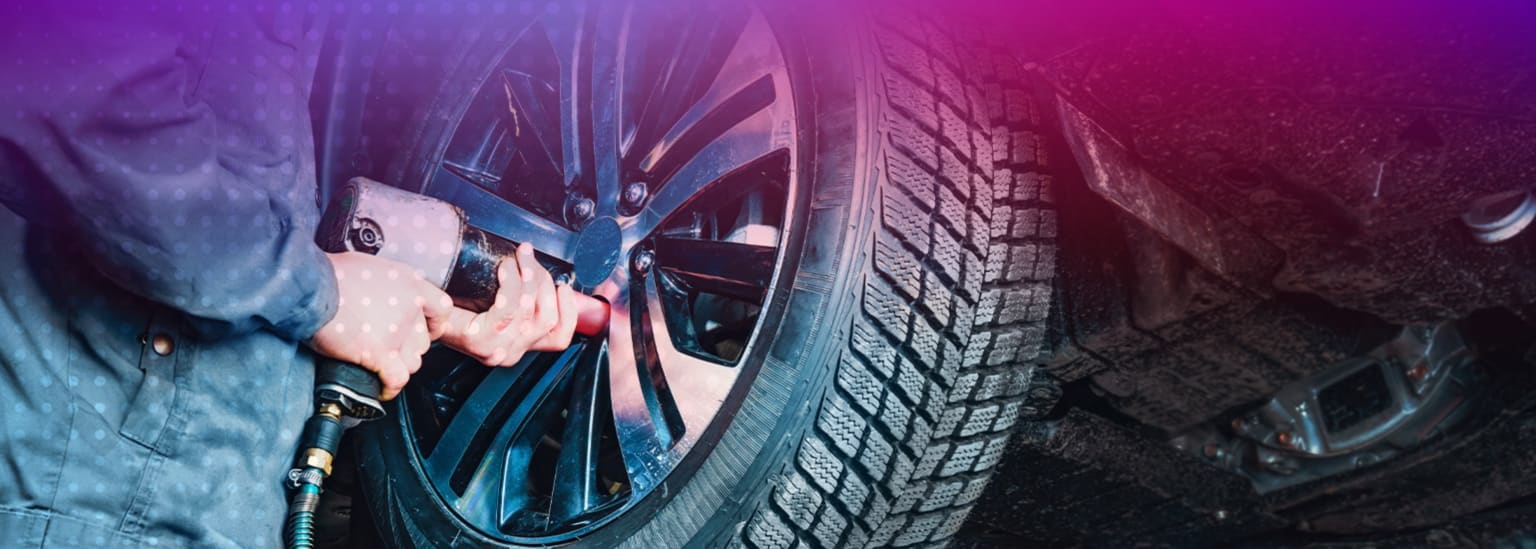 A mechanic using an impact wrench to tighten lug nuts on a car's wheel, with the underside of the car visible in the background.