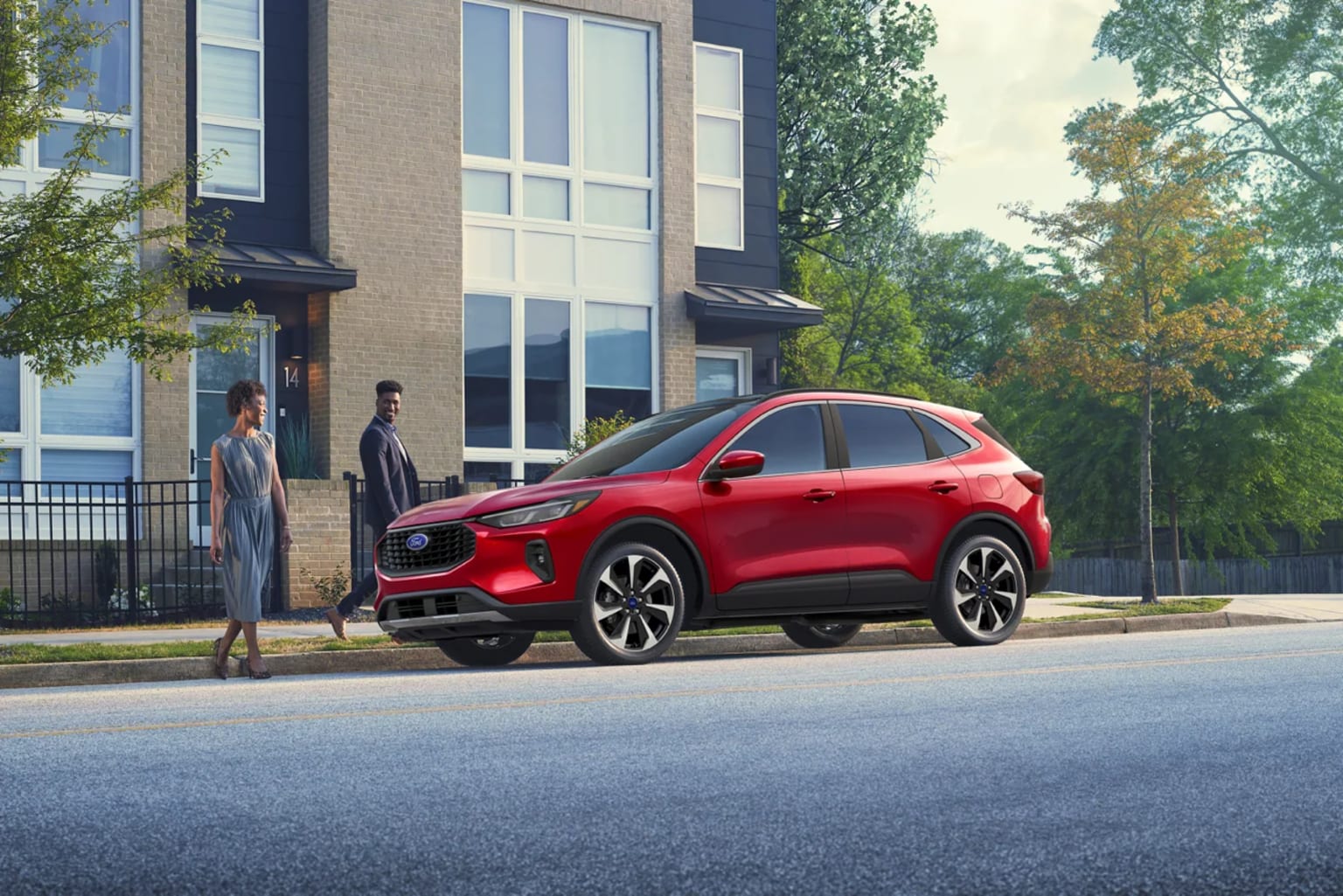 A red SUV parked on the street in front of a modern multi-story building, surrounded by trees and pedestrians walking on the sidewalk.