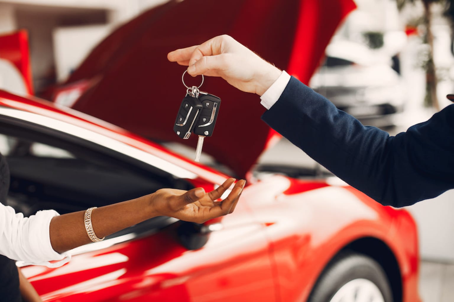 a woman handing a car key to a man in a suit and tie in front of a red sports car