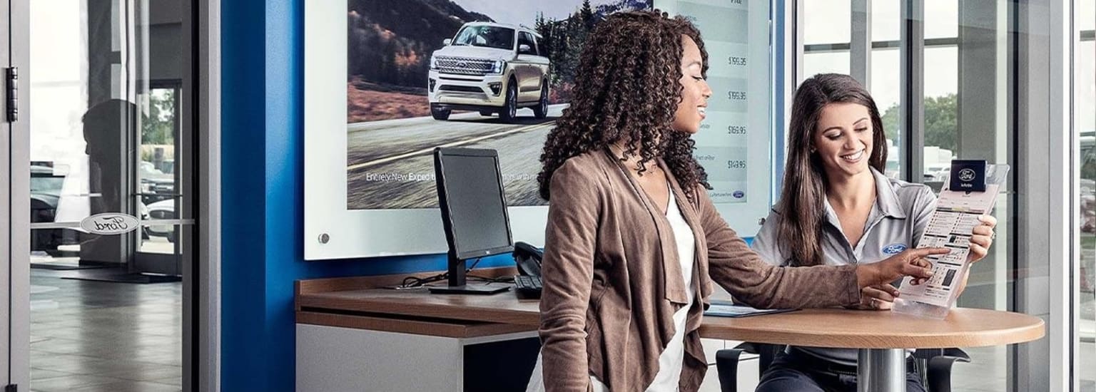 Two women sit at a desk, one with curly hair and the other with straight hair, in front of a display showing an image of a large vehicle.