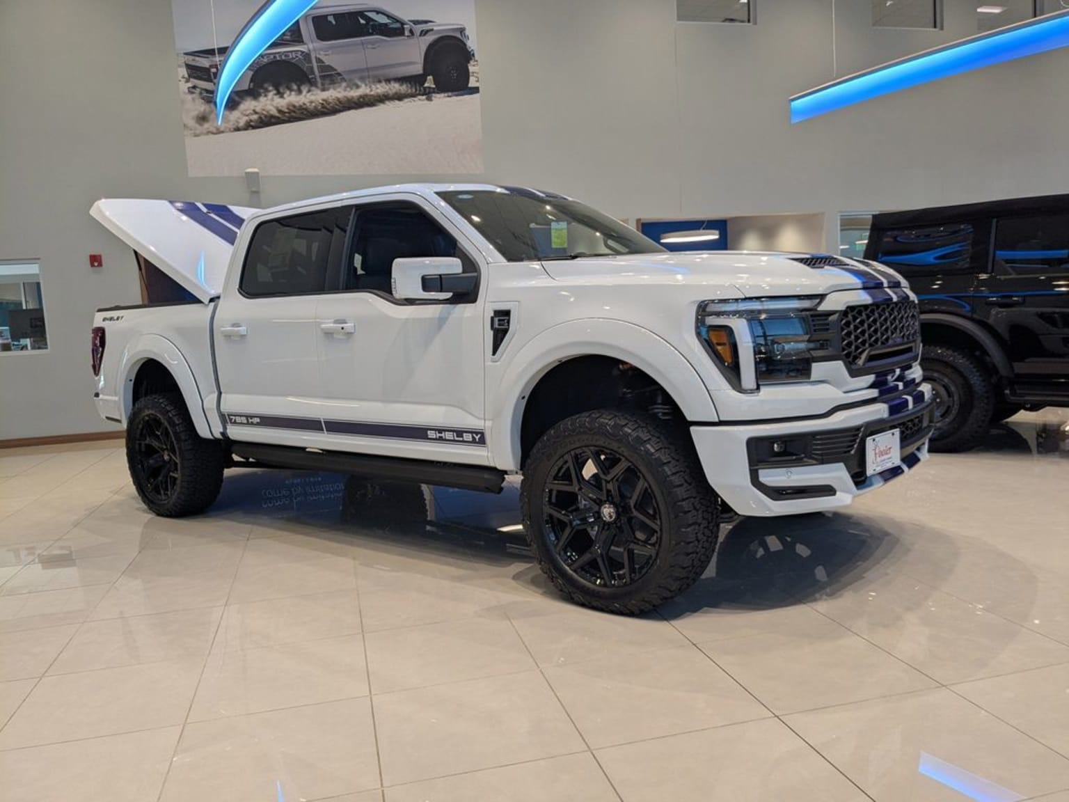 A large, white pickup truck with off-road modifications, including large tires and a lifted suspension, is displayed in a showroom setting with a background image of a similar truck in an outdoor environment.
