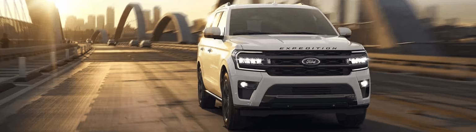 A large, white SUV with the "Expedition" branding on the front grille, parked on a wooden dock or pier against a backdrop of blurred trees and foliage.