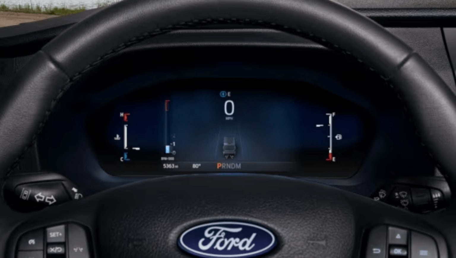 Close-up of a car's digital dashboard displaying speed and other information, with the logo visible on the steering wheel in the foreground and a blurred exterior in the background.
