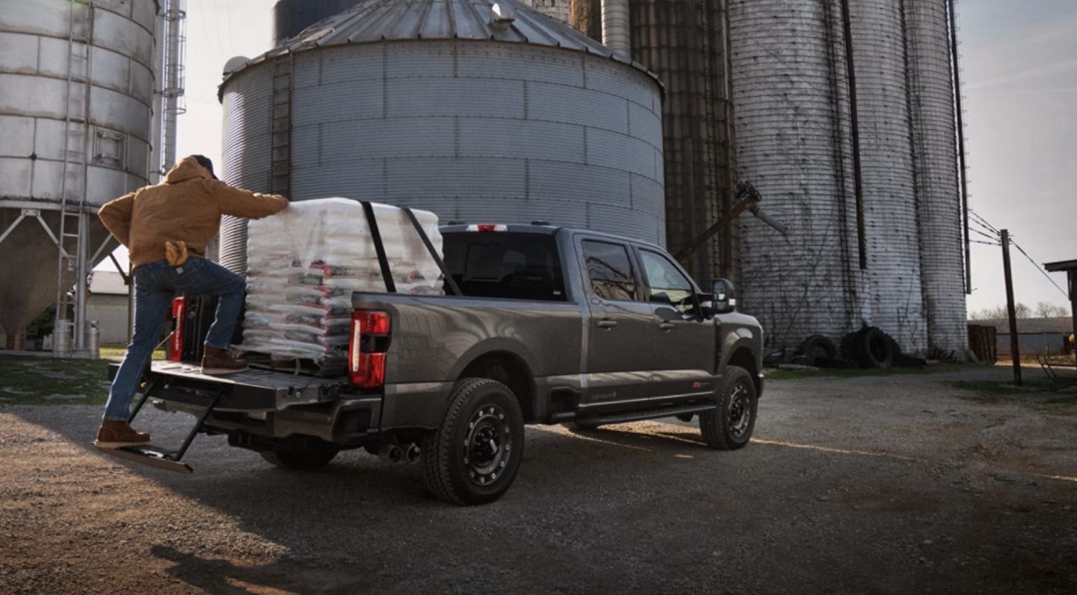A man loads white bags into the bed of a grey pickup truck parked in front of large industrial silos.