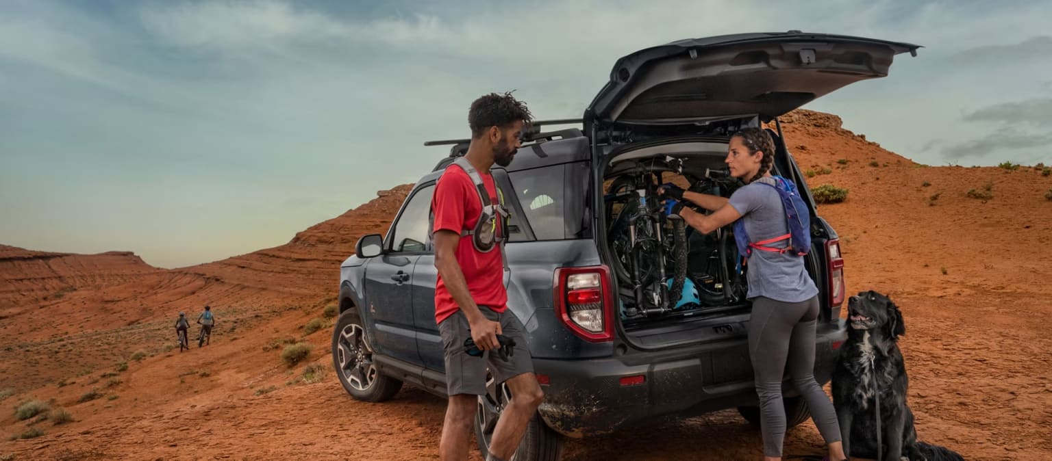 Group of people standing next to an open-trunk vehicle in a rugged, desert-like landscape with reddish-brown rock formations in the background.